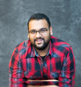 Sunny Singh, owner of SSinghPhoto, wearing a red plaid shirt and glasses, smiling against a textured gray backdrop