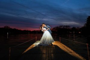 Bride and groom sharing a romantic kiss on a wooden dock at sunset, with vibrant purple and orange skies reflecting over the water.
