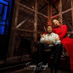 Engaged couple sitting on a leather chair in a moody, stylish studio with rustic wood backdrop and bold red and white outfits