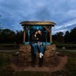 Engaged couple sitting together on a stone wishing well in a park at dusk, smiling and cuddling