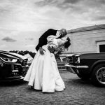 Bride and groom share a dramatic kiss between two muscle cars in a black-and-white wedding photo