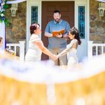 Two LGBTQ brides hold hands and exchange vows during their outdoor wedding ceremony with guests and florals in view