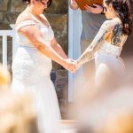 Two LGBTQ brides hold hands during their outdoor wedding ceremony while exchanging vows, captured from the guests' perspective