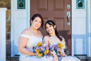 LGBTQ brides sitting together on a front porch in wedding dresses, smiling and holding colorful floral bouquets